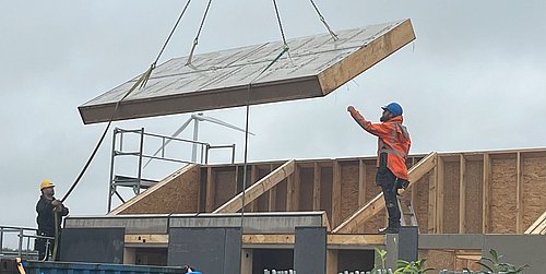 Foto, zwei Bauarbeiter mit Schutzhelmen und Warnkleidung heben mit einem Kran ein großes, rechteckiges Bauteil über einem Rohbau an.