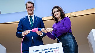 Foto, zwei Personen stecken symbolisch zwei Stecker zusammen, an denen das Logo des Gemeinschaftswerks Nachhaltigkeit befestigt ist.