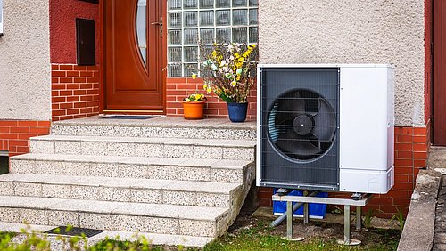Foto, Außenbereich eines Hauses mit Treppe, brauner Haustür, Blumendekoration auf der Treppe und einer Wärmepumpe auf dem Rasen.