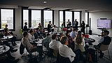 Foto, Mehrere Personen sitzen in einem modernen Konferenzraum und hören einem Vortrag vor einem Bildschirm zu.