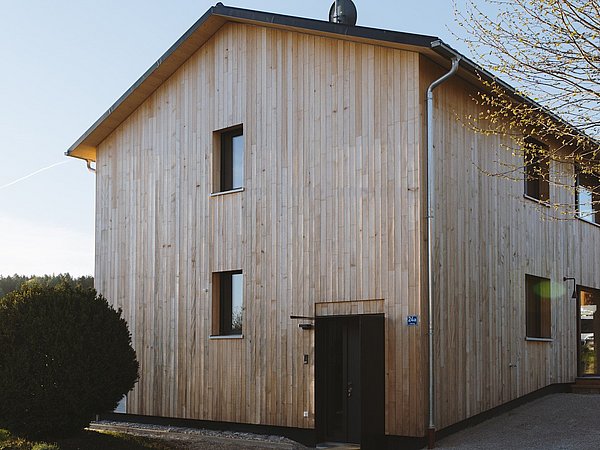 Haus mit Fassade aus vertikal verlaufenden Holzlatten, rechteckige Fenster, Eingangstür, Baum und Hecke im Vordergrund.