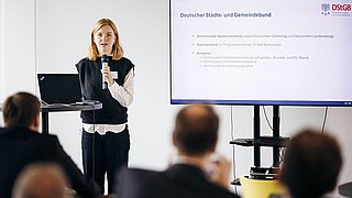 Foto, Frau mit Mikrofon und Laptop hält Vortrag vor Publikum, rechts Bildschirm mit Präsentationsfolie zum Deutschen Städte- und Gemeindebund.