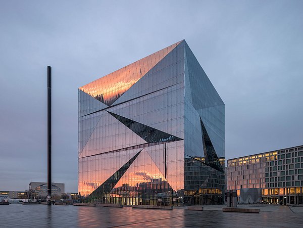 Foto, Außenansicht von einem mehrstöckigen, verspiegelten, quadratischen Gebäudes. In er Fassade spiegeln sich weitere Gebäude und der rötliche Himmel.