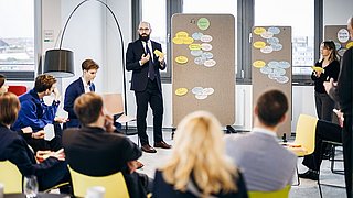 Foto, Mann im Anzug steht vor Pinnwänden mit bunten Notizzetteln, umgeben von sitzenden und stehenden Personen in modernem Büro.