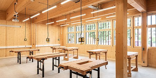 Foto, großer Werkraum mit Holzwänden und Holzdecke. In dem Raum befinden sich mehrere Werkbänke, die in Reihen stehen.