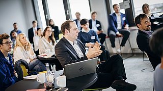 Foto, Mann in Anzug sitzt an Tisch mit Laptop, gestikuliert mit den Händen, im Hintergrund mehrere sitzende Personen in Businesskleidung.