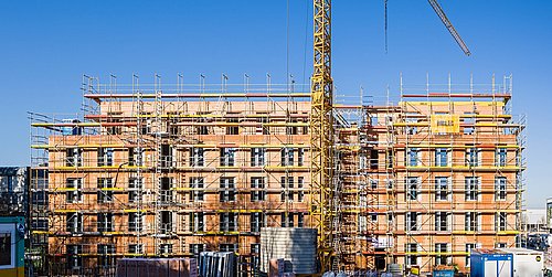 Foto, Baustelle eines mehrstöckigen Gebäudes. Das Gebäude ist eingerüstet, im Vordergrund steht ein Kran.