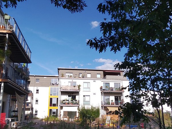 Foto, Im Vordergrund stehen mehrere Bäume mit dichtem Laub, die einen Teil des Bildes beschatten. Dahinter sind zwei moderne Wohnhäuser mit mehreren Balkonen zu sehen. Die Fassaden sind weiß und grau, einzelne Elemente sind gelb hervorgehoben. Auf den Balkonen befinden sich Pflanzen und Möbel. Der Himmel ist klar und blau, nur wenige kleine Wolken sind sichtbar.