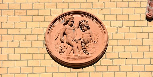 Foto, Fassadenstuck aus Terracotta mit Putten an einer gelben Klinkerfassade.