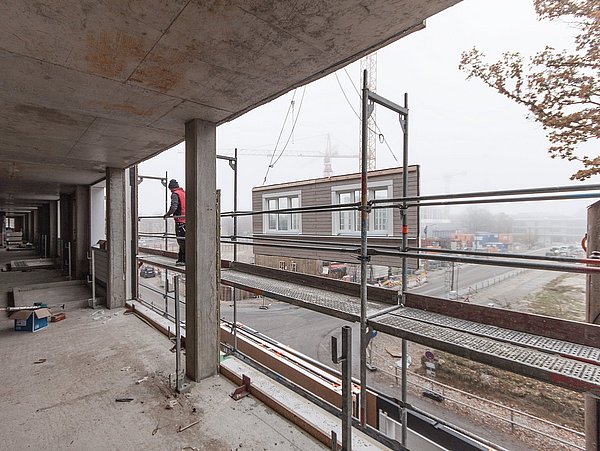 Foto, Ausblick von Baustelle eines Rohbaus. Vor dem Gebäude steht ein Gerüst. An einem Kran wird ein Fassadenelement mit Fenster montiert.