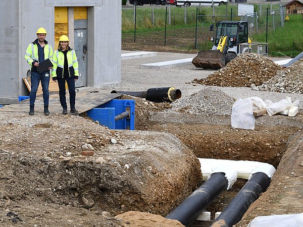 Zwei Mitarbeitende mit Schutzhelmen und gelben Warnwesten stehen neben einer Baustelle mit großen Rohrleitungen und Erdarbeiten für die Anlieferung einer Energiezentrale.