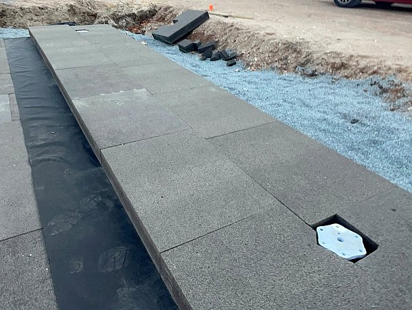 Foto, eine teilweise fertiggestellte Baustelle mit grauen Bodenplatten eines Fundamentes, umgeben von losem Kies und Geräten.