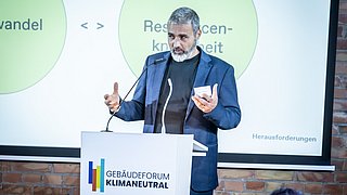 Foto, Mann in dunklem Anzug spricht am Rednerpult mit Logo 'Gebäudeforum Klimaneutral', im Hintergrund Präsentationsfolie mit den Begriffen 'Resilienz' und 'Herausforderungen'.