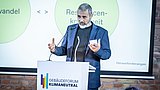 Foto, Mann in dunklem Anzug spricht am Rednerpult mit Logo 'Gebäudeforum Klimaneutral', im Hintergrund Präsentationsfolie mit den Begriffen 'Resilienz' und 'Herausforderungen'.