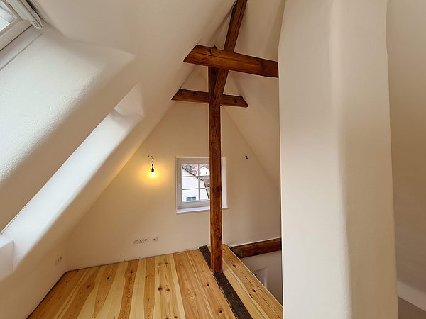 Foto, Ausgebautes Dachgeschoss mit sichtbaren Holzbalken, hellem Holzboden und kleinem Fenster