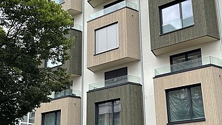 Foto, modernes Mehrfamilienhaus mit Balkonen mit Glasgeländern und abwechselnden Holzstrukturen, umrahmt von üppigen grünen Bäumen.