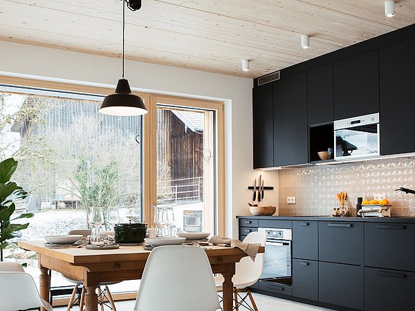Moderne Küche mit schwarzen Schränken, Holztisch und weißen Stühlen vor großen Fenstern mit Blick auf verschneite Landschaft.