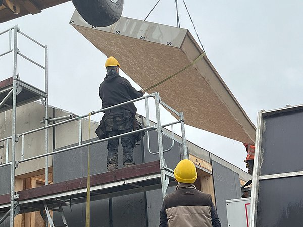 Foto, zwei Bauarbeiter mit gelben Schutzhelmen montieren auf einem Gerüst ein großes Holzpaneel an einem Hausdach.