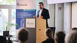 Foto, Mann steht hinter einem Rednerpult mit Mikrofon vor einem Banner mit dem Schriftzug "Gebäudeforum klimaneutral" und spricht vor Publikum.