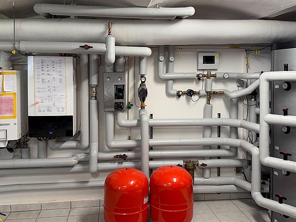 Foto, Heizungsanlage: Mehrere isolierte Rohrleitungen verlaufen horizontal und vertikal an einer weißen Wand. Zwei rote, zylindrische Druckbehälter stehen nebeneinander auf dem gefliesten Boden. Rechts befindet sich ein großer, silberfarbener Speicherbehälter mit mehreren angeschlossenen Rohren. An der Wand sind verschiedene technische Geräte, Schaltkästen, Anzeigen und ein Wandthermostat montiert. Zwei große, übersichtliche Schaltpläne sind sichtbar angebracht.