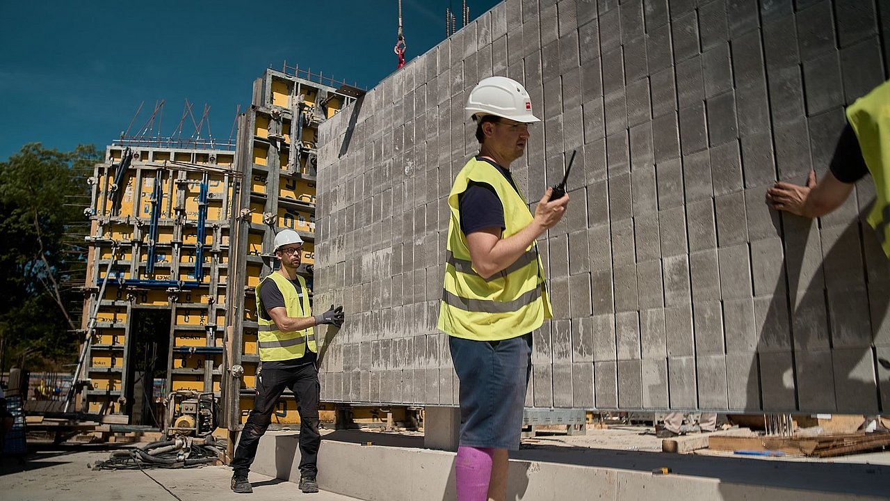 Foto, drei Bauarbeiter in Sicherheitswesten und Helmen auf einer Baustelle vor einer Mauerwand, einer hält ein Funkgerät.