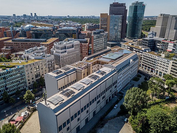 Foto, Luftaufnahme vom Potsdamer Platz in Berlin mit einem langen, rechteckigen Gebäude, der Kältezentrale der Berliner Energie und Wärme GmbH im Vordergrund, umgeben von modernen Bürogebäuden. Im Hintergrund sind die beiden Hochhäuser der Deutschen Bahn und des Kollhoff Towers, das Ritz Hotel sowie weitere Gebäude sichtbar.