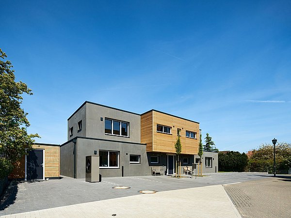 Foto, Modernes zweistöckiges Wohnhaus mit grauer und holzverkleideter Fassade, großen Fenstern und gepflastertem Hof unter blauem Himmel