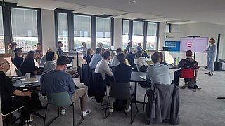 Foto, Mehrere Personen sitzen in einem hellen Konferenzraum und schauen auf eine Präsentation auf einem Bildschirm