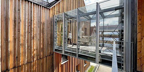 Foto, ein Verbindungssteg im dritten Stockwerk aus Metall und Glas verbindet zwei Holzgebäude.