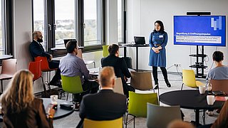 Foto, Frau im blauen Kleid steht vor Bildschirm mit Text "Begrüßung & Eröffnungsimpulse", Publikum sitzt auf bunten Stühlen in modernem Raum.
