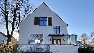 Saniertes Haus mit weißer Fassade, braunen Fensterläden und großen Fenstern, umgeben von kahlen Bäumen und niedrigem Zaun.