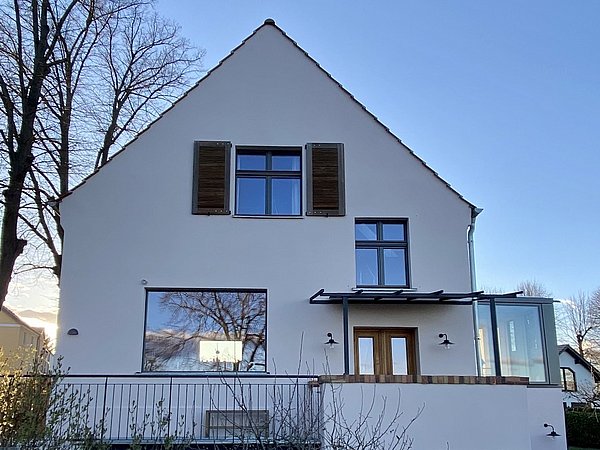 Saniertes Haus mit weißer Fassade, braunen Fensterläden und großen Fenstern, umgeben von kahlen Bäumen und niedrigem Zaun.