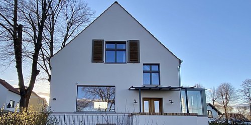 Saniertes Haus mit weißer Fassade, braunen Fensterläden und großen Fenstern, umgeben von kahlen Bäumen und niedrigem Zaun.