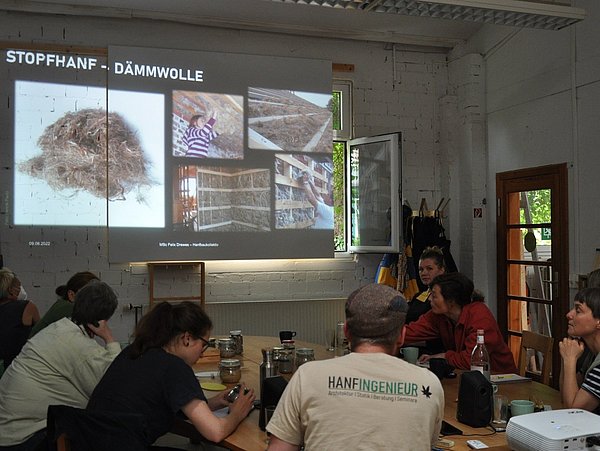 Foto, im Vordergrund sitzen mehrere Personen an einem Tisch. Im Hintergrund wird eine Präsentation an die Wand projiziert. 