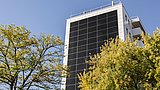 Foto, ein hohes, modernes Gebäude mit Sonnenkollektoren an der Fassade, umgeben von Bäumen unter einem klaren blauen Himmel.