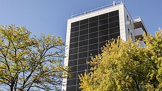 Foto, ein hohes, modernes Gebäude mit Sonnenkollektoren an der Fassade, umgeben von Bäumen unter einem klaren blauen Himmel.