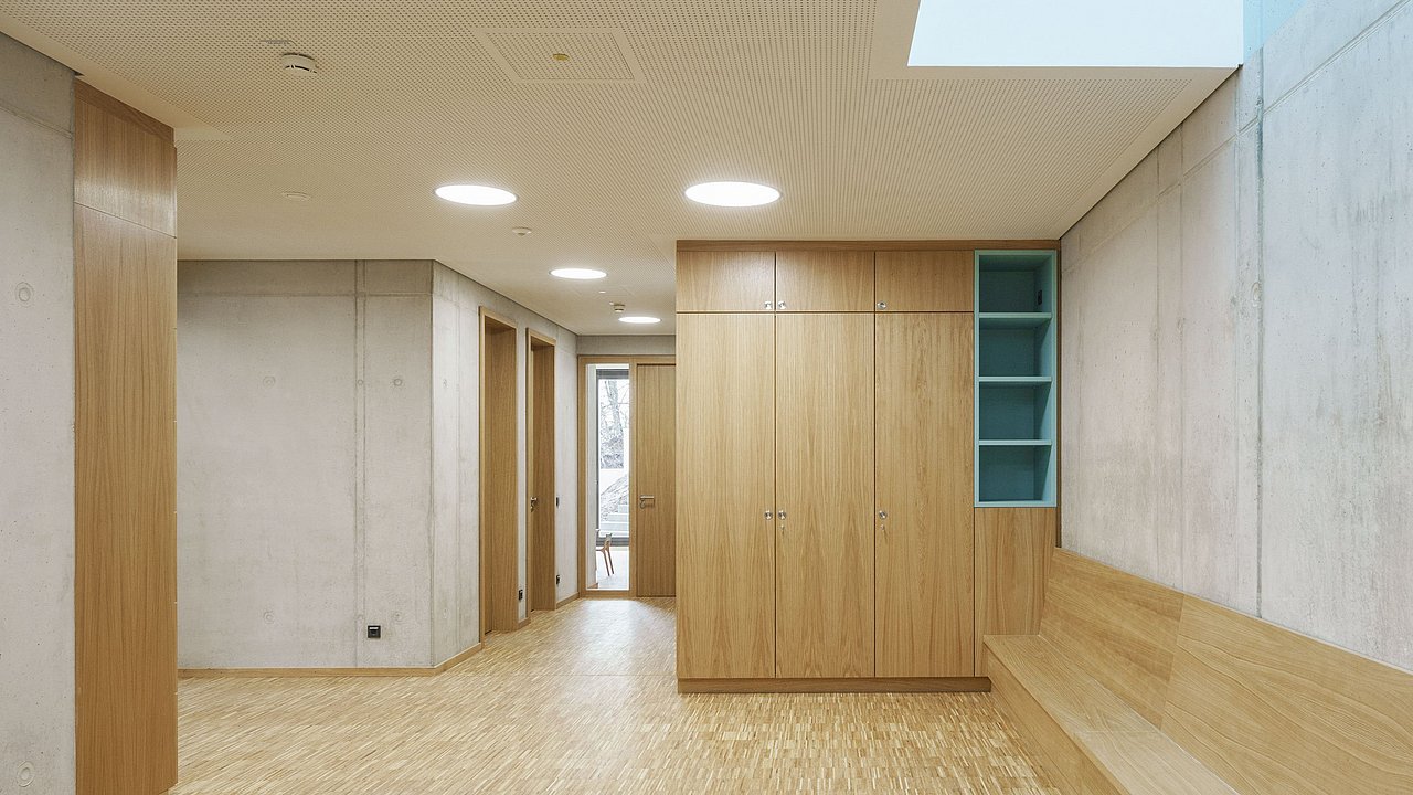 Foto, Flur mit Betonwänden und Holzboden. Rechts befindet sich ein Schrank und eine lange Sitzbank.