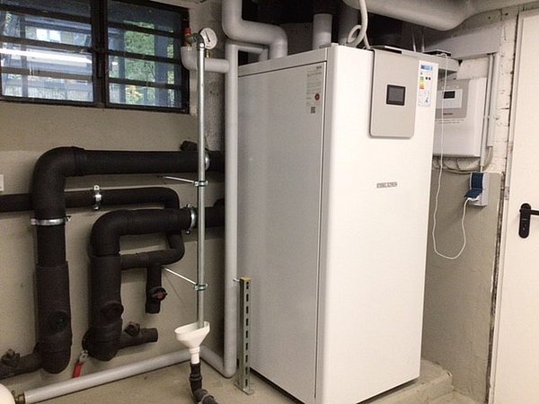 Foto, Weiße Heizungsanlage mit angeschlossenen schwarzen und weißen Rohrleitungen in einem Kellerraum