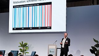 Foto, ein Redner hält einen Vortrag auf einer Bühne und zeigt Präsentationsfolien.