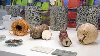 Foto, auf einem Tisch liegen aufgereiht verschiedene bio-basierte Materialien und bio-basierte Reststoffe, die im Betonbau einsetzbar sind. Im Hintergrund sind Muster-Vasen verschiedener Betonarten zu sehen.