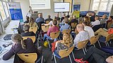 Foto, vortragende Person steht vor Publikum in einem Seminarraum mit Präsentationsbildschirm und Rollup im Hintergrund