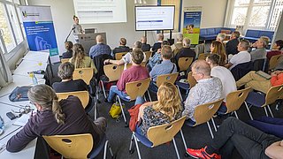 Foto, vortragende Person steht vor Publikum in einem Seminarraum mit Präsentationsbildschirm und Rollup im Hintergrund