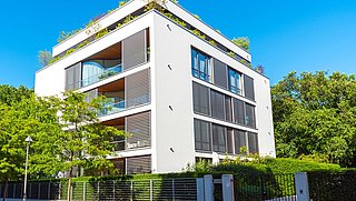 Foto, Modernes, weißes Wohngebäude mit großen Fenstern, Balkonen und begrüntem Dach in sonniger Umgebung