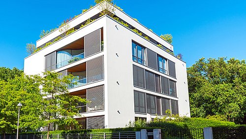 Foto, Modernes, weißes Wohngebäude mit großen Fenstern, Balkonen und begrüntem Dach in sonniger Umgebung