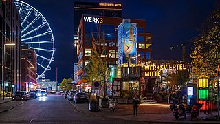 Foto, Nachtaufnahme des Werksviertel Mitte in München mit beleuchtetem Riesenrad links und Gebäuden rechts