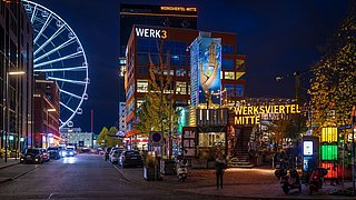 Foto, Nachtaufnahme des Werksviertel Mitte in München mit beleuchtetem Riesenrad links und Gebäuden rechts