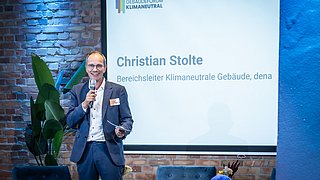 Foto, Christian Stolte hält eine Rede bei der Jahrestagung 2024 des Gebäudeforums Klimaneutral, er ist Bereichsleiter Klimaneutrale Gebäude bei dena, steht vor einer Leinwand mit seinem Namen und Titel.