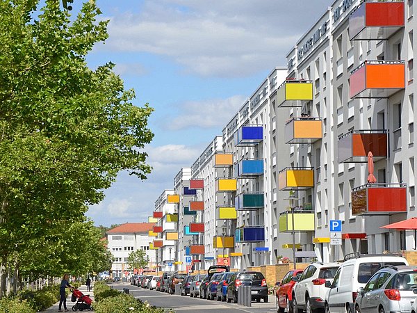 Foto, Straßenszene mit mehrstöckigen Wohnhäusern, deren Balkone in verschiedenen Farben, vielen grünen Bäumen links, und parkenden Autos entlang der Straße