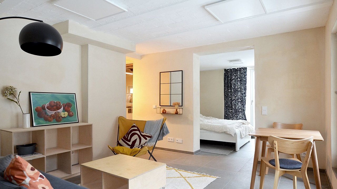 Foto, Wohnzimmer mit grauem Sofa, Holzregal, Sessel mit buntem Kissen, Holztisch auf Rollen, Blick ins Schlafzimmer mit weißem Bett und dunklem Vorhang