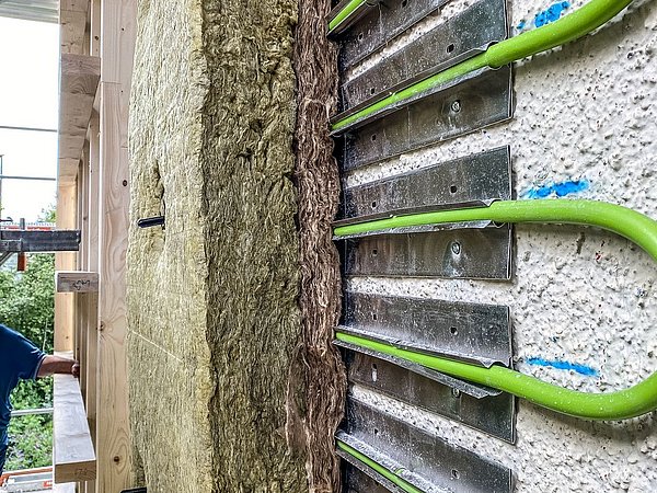 Foto, Nahaufnahme von dünnen grünen Rohren, die an einer Wand mit Metallhaltern befestigt sind, neben einer Schicht Dämmmaterial